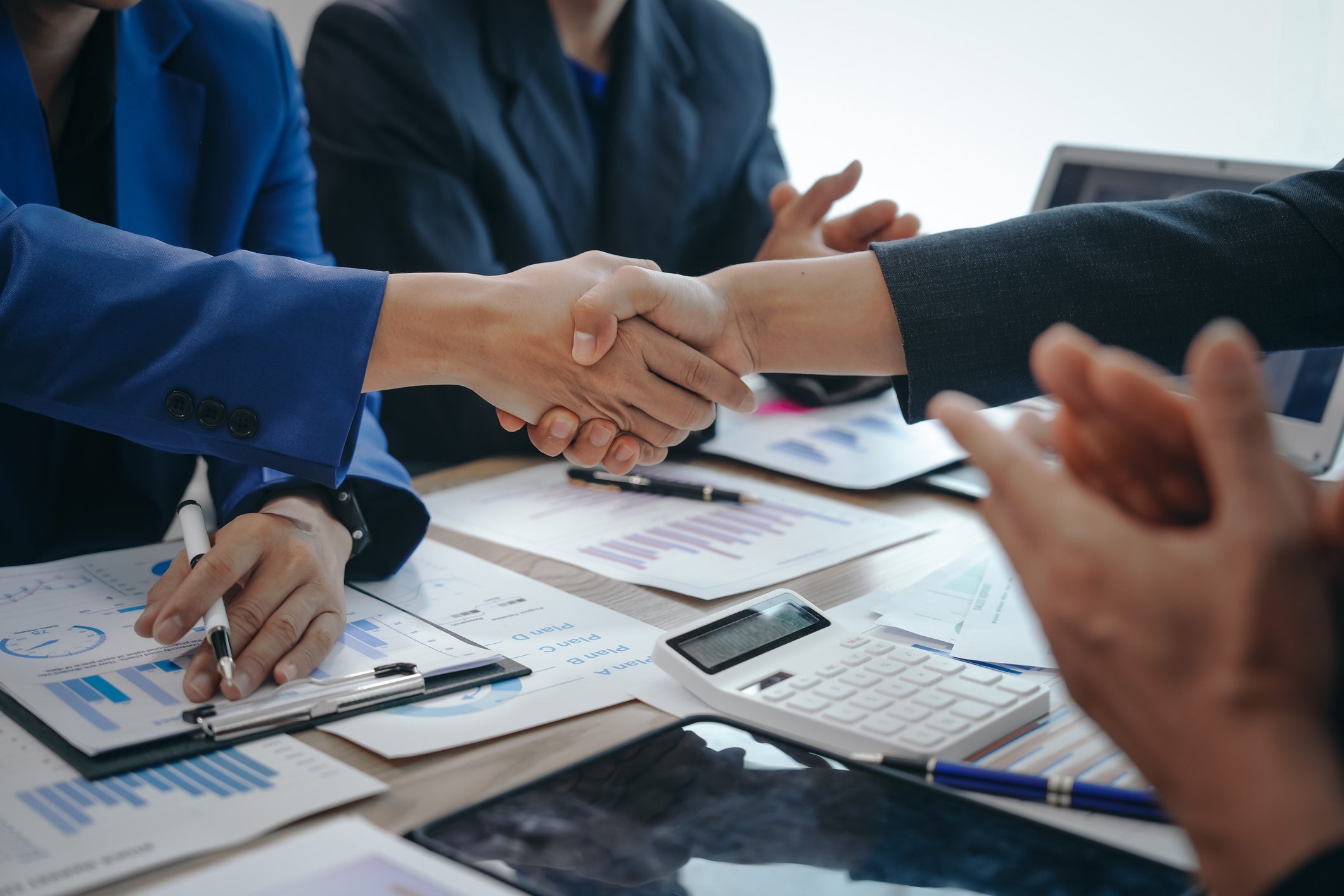 Hands shaking in a business meeting with financial papers, calculator, and laptop