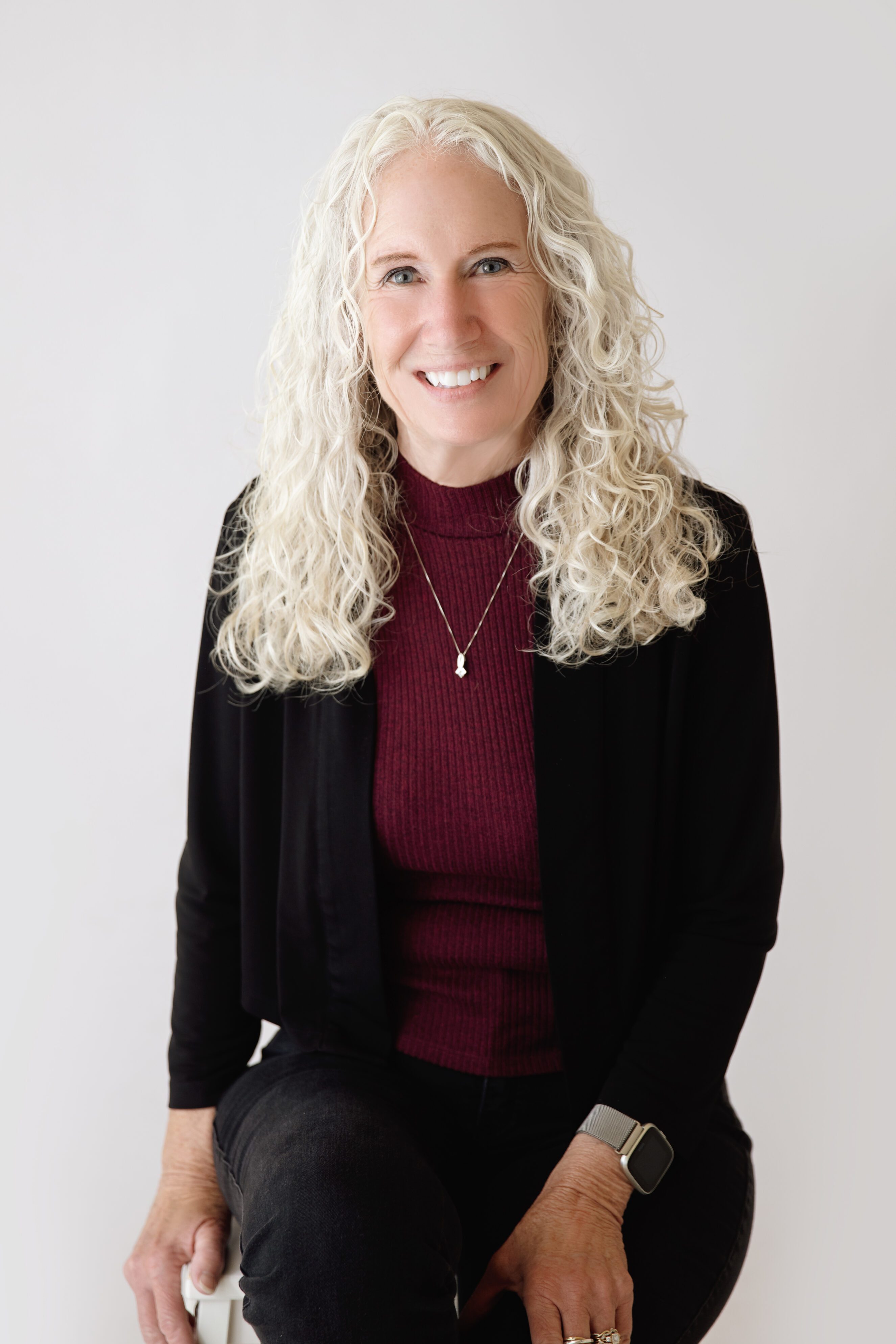 Smiling woman with long curly white hair, wearing burgundy shirt, black cardigan,