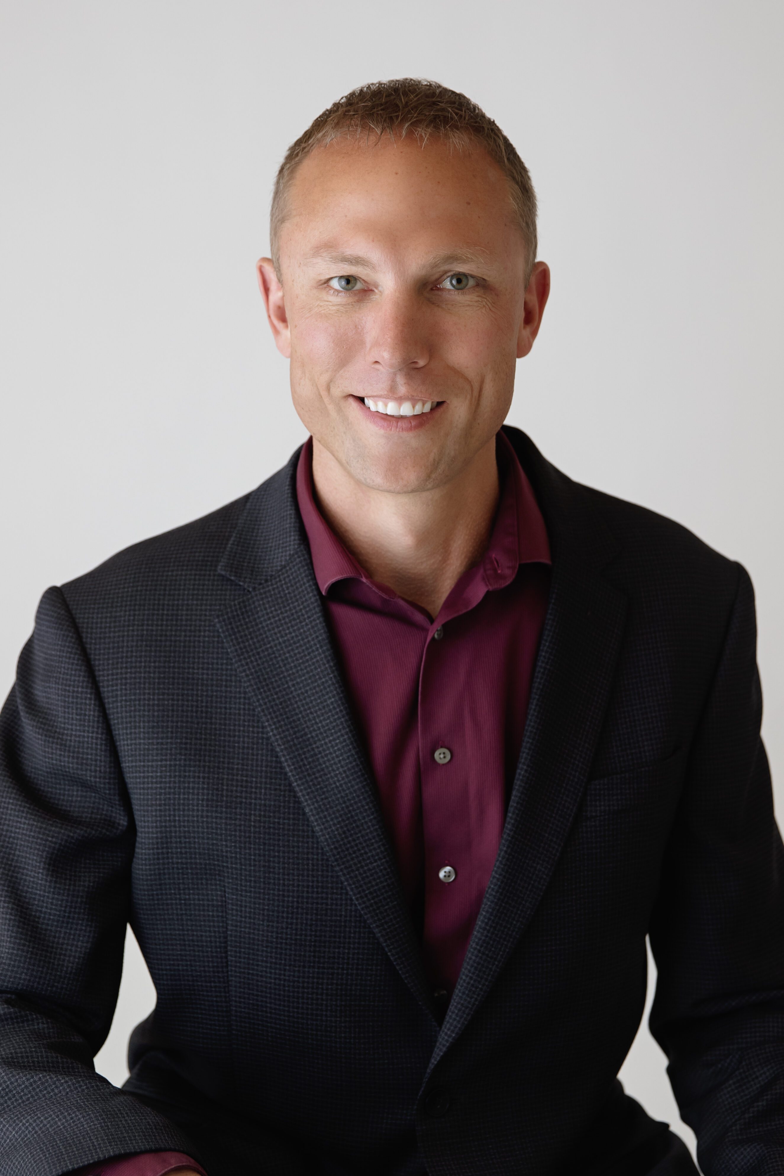 Man wearing dark checkered blazer and maroon shirt smiling against plain background