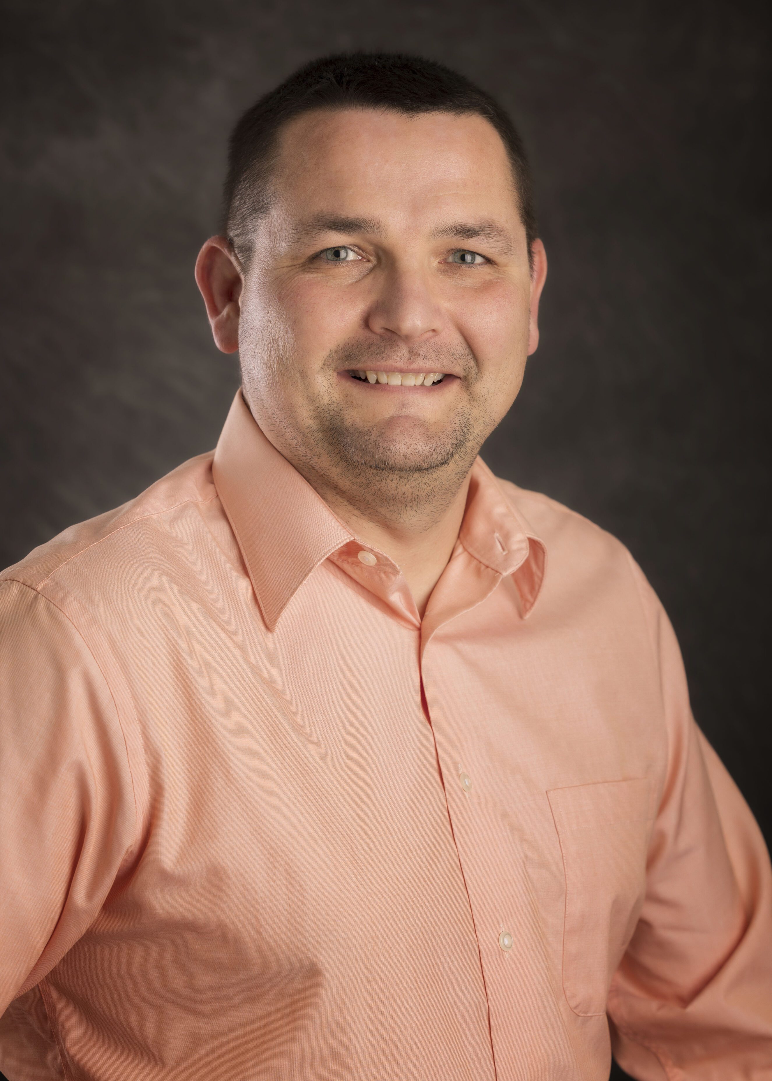 Man with short dark hair wearing a peach button-up shirt smiling against